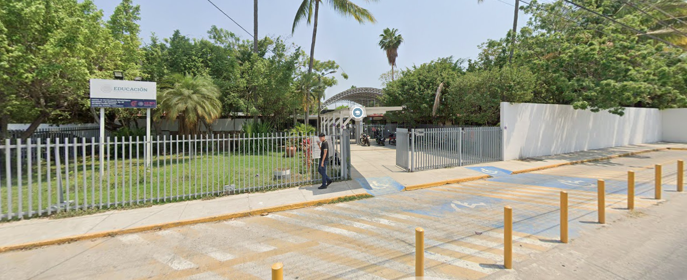Fotografía en la que se muestra una rampa que lleva a una reja de color gris por la que se puede ingresar al plantel. Las instalaciones están rodeadas por rejas de color gris y muros blancos. A la izquierda y a la derecha de la imagen hay áreas verdes.